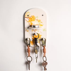 Könnte beinhalten: Ein cremefarbener Keramik-Schlüsselhalter mit einem floralen Design in Gelb und Orange. Er verfügt über ein kleines Regal mit einer brennenden Kerze und drei Schlüsselhaken mit Leder- und Metall-Schlüsselanhängern. Der Schlüsselhalter ist an einer weißen Wand befestigt.