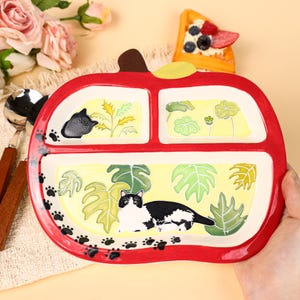 May include: A red, apple-shaped divided plate with a whimsical cat design. The plate features a black and white cat illustration, paw prints, and leaf and floral accents. The plate is divided into three sections, perfect for serving a child's meal.