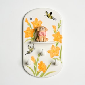 May include: A decorative key holder with a white background, featuring a painted design of orange flowers, green leaves, and yellow butterflies. A small dog figurine with a pink bow sits on a shelf above three white hooks.
