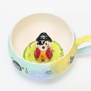 Könnte beinhalten: Eine Keramiktasse mit einer Piratenhund-Figur im Inneren. Der Hund trägt einen schwarzen Hut mit Totenkopf, eine Augenklappe und ein gelb-rotes Outfit. Die Tasse hat eine farbenfrohe Außenseite und einen Henkel.