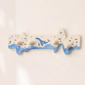 May include: A wall-mounted hook rack with an irregular, wavy top and bottom edge. The off-white surface features a painted blue whale, small fish, and star motifs. The base includes four protruding wooden pegs and two visible mounting screws.