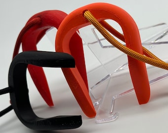 Könnte beinhalten: Drei U-förmige Objekte in Rot, Orange und Schwarz, jeweils mit einer Schnur befestigt. Das orangefarbene Objekt hat eine beige Schnur. Die Objekte befinden sich auf einer weißen Oberfläche, mit einem transparenten Kunststoffständer im Hintergrund.