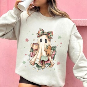 Könnte beinhalten: Ein beiges Sweatshirt mit Rundhalsausschnitt und einer Grafik eines Geistes mit floraler Schleife, der eine Tasse und einen Lebkuchen hält. Der Geist ist in eine karierte Decke gehüllt und von kleinen grünen und roten Schneeflocken umgeben.