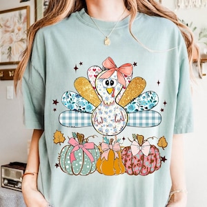 Könnte beinhalten: Ein hellgrünes T-Shirt mit einer Grafik eines Truthahns mit rosa Schleife und gemusterten Schwanzfedern, platziert über drei Kürbissen, die mit Schleifen und verschiedenen Mustern wie Punkten, Leopardenmuster und einfarbigem Orange verziert sind.