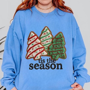 Könnte beinhalten: Ein hellblaues Sweatshirt mit Rundhalsausschnitt und einem zentralen Motiv aus drei stilisierten Weihnachtsbäumen mit gestrickten Texturen in Rot, Weiß und Grün. Unter den Bäumen steht der Text "tis the season" in Schwarz.