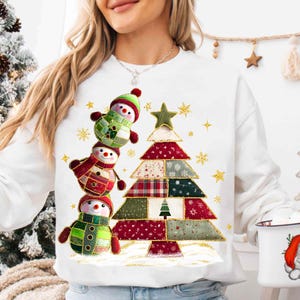 Könnte beinhalten: Ein weißes Langarm-Sweatshirt mit einem Grafikdruck von drei gestapelten Schneemännern und einem Weihnachtsbaum im Patchwork-Stil. Die Schneemänner und der Baum sind mit verschiedenen roten, grünen und weißen Mustern sowie kleinen goldenen Sternen verziert.