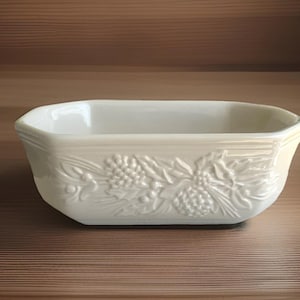 May include: Off-white rectangular ceramic baking dish with embossed grape and leaf design. The dish has a slightly flared rim and is set against a wooden background. A classic piece for baking and serving.
