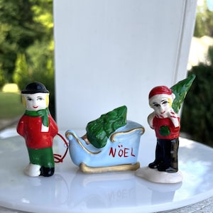 May include: A set of three Christmas figurines. A figurine of a person in a red coat, green trousers, and a black hat and scarf. A light blue sleigh with a green tree and the word "NOEL". A figurine of a person holding a green tree.