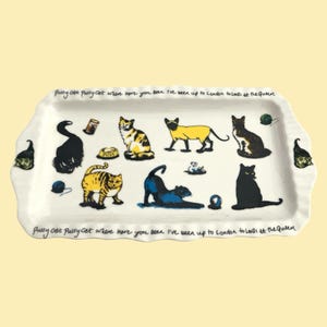 Könnte beinhalten: Ein rechteckiges weißes Keramiktablett mit gewelltem Rand, verziert mit Illustrationen verschiedener Katzen in Schwarz, Gelb und Braun. Der Text Pussy cat Pussy cat where have you been I've been up to London to look at the Queen steht am oberen und unteren Rand.