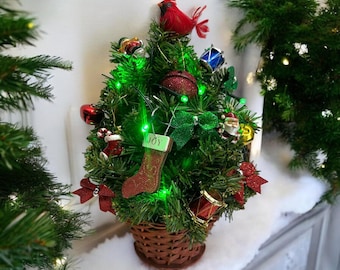 May include: A small Christmas tree in a wicker basket, decorated with red and green baubles, a red cardinal, and green lights. A stocking with the word "JOY" hangs from the tree. The tree is on a white surface.