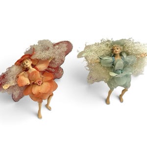 May include: Two decorative fairy figurines with outstretched arms. One figure wears an orange petal-like outfit with matching wings and white curly hair. The other figure wears a light blue petal-like outfit with matching wings and white curly hair.