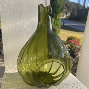 Może przedstawiać: Oliwkowozielony szklany wazon o pękatym korpusie i rozkloszowanej, karbowanej górze. Wazon ma okrągły otwór z boku i pionowe żłobienia. Wazon stoi na białej powierzchni.