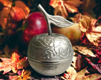 Könnte beinhalten: Ein silberner Apfel aus Metall mit einem detaillierten Blumenmuster. Der Apfel liegt auf einem Bett aus Herbstblättern.