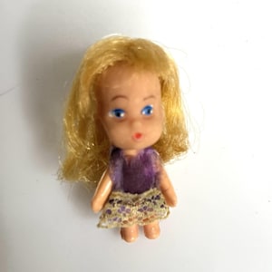 May include: A small doll with blonde hair, blue eyes, and a painted red mouth. The doll wears a purple sleeveless top paired with a short, beige, lace-trimmed skirt featuring a delicate floral pattern. The figure has a light skin tone.