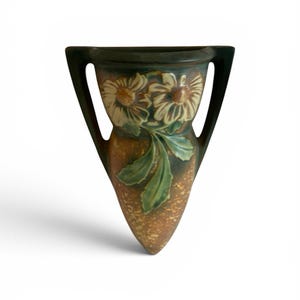 May include: A decorative vase with a dark green rim and handles, featuring a floral design with cream-colored flowers and green leaves. The vase has a speckled brown body and a unique, elongated shape.