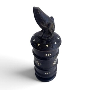 May include: A dark brown cylindrical wooden container with a stacked design and a carved frog figure on the lid. The exterior is decorated with small, inlaid white triangular and diamond-shaped pieces arranged in geometric patterns around the body.