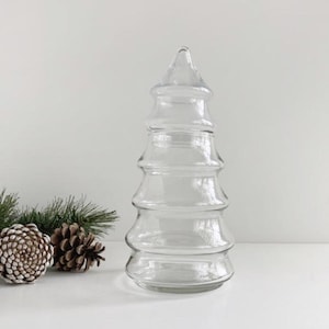 Puede incluir: Un jarrón decorativo transparente con forma de árbol de Navidad. El jarrón tiene un diseño escalonado y una parte superior puntiaguda. El jarrón está sobre una superficie blanca con agujas de pino y piñas en el fondo.