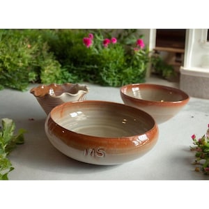 Puede incluir: Tres cuencos de cerámica de diferentes tamaños. El cuenco más grande es blanco con un borde marrón y las letras "MS" en la parte inferior. El cuenco mediano es marrón con un interior blanco. El cuenco más pequeño es blanco con un borde rizado.