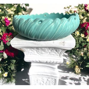 May include: A teal blue ceramic planter with a wavy design and drainage holes. The planter is sitting on a white pedestal with ornate details.