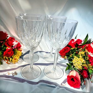 May include: Four clear glass wine glasses with a cut crystal design. The glasses are arranged in a square shape on a white tablecloth with a bouquet of red and yellow flowers in the background.