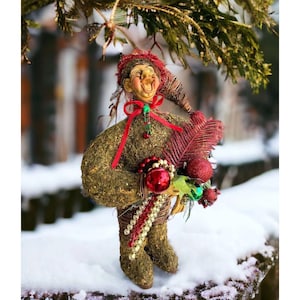 May include: A whimsical Christmas ornament featuring a mossy green elf with a red hat and a red bow. The elf is holding a sprig of red and green foliage and a red ball.