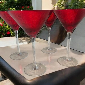 Könnte beinhalten: Vier rote Martinigläser mit klaren Stielen und Böden. Die Gläser haben ein strukturiertes Muster und sind auf einer hellen Oberfläche angeordnet. Der Hintergrund umfasst Grün und einen Blick auf eine Terrasse.