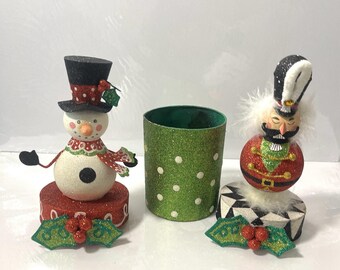 May include: Festive Christmas decorations featuring a snowman, a green polka-dot cylinder, and a nutcracker. The snowman has a black top hat and a red scarf. The nutcracker is in a red and green uniform. Each sits on a decorative base.