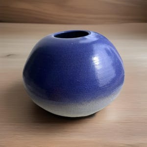 May include: A round, blue ceramic vase with a wide opening. The vase has a smooth, glossy finish and a gradient color that fades from a deep blue to a light gray at the base. The vase sits on a light brown wooden surface.