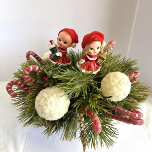 Peut inclure: Une décoration de Noël festive avec deux figurines vintage en robes et chapeaux rouges, nichées parmi des aiguilles de pin vertes, des cannes en sucre et des boules décoratives blanches. L'arrangement évoque un thème de vacances nostalgique.