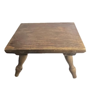 May include: A small, square wooden table with a distressed finish. The table has four curved legs and a light brown color. The tabletop is approximately 18 inches square.