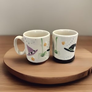 May include: Two white ceramic Halloween-themed mugs. One mug has a handle and a purple witch hat design, the other has a black witch hat design. Both mugs feature green spiders and candy corn accents.