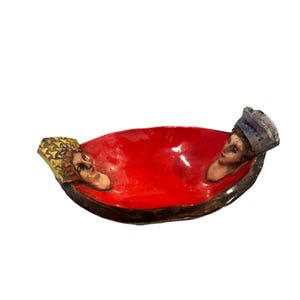Może przedstawiać: Czerwona ceramiczna miska z dwoma twarzami wyrzeźbionymi po bokach. Twarze są pomalowane szczegółowo i mają kapryśny wyraz.