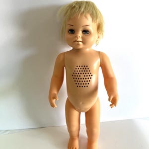Pode incluir: Uma boneca de plástico com cabelo loiro curto e olhos azuis. A boneca tem um acabamento de pele cor de pêssego e uma grade de alto-falante hexagonal com numerosas pequenas perfurações circulares no centro do tronco.