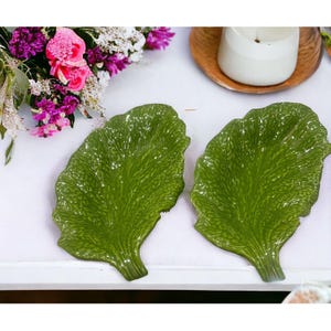Könnte beinhalten: Zwei grüne, blattförmige Keramikplatten mit strukturierten Details. Sie liegen auf einer weißen Oberfläche, mit einem Strauß rosa und lila Blumen und einer weißen Kerze im Hintergrund. Die Teller sind so gestaltet, dass sie Salatblättern ähneln.