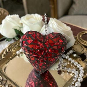May include: A red mosaic heart ornament with a silver chain for hanging. The heart is made of small, irregularly shaped red glass pieces. It sits on a gold-colored tray with white roses and a pearl necklace in the background.