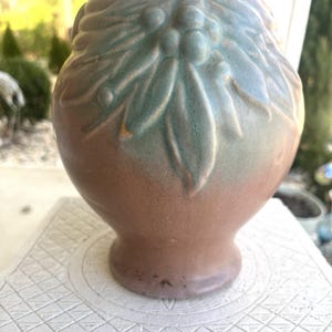 Könnte beinhalten: Eine Keramikvase mit rundem Körper und ausgestelltem Rand. Die Vase ist hauptsächlich braun mit grünen und blauen Akzenten auf dem oberen Teil, mit einem Blatt- und Beerenmuster. Die Vase steht auf einer weißen Oberfläche.