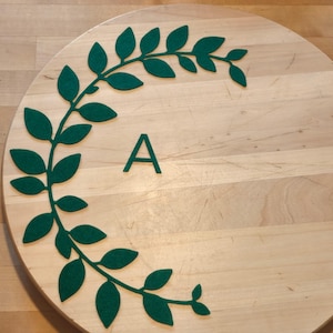 Peut inclure: Un rondin de bois avec une guirlande de feuilles en feutre vert et la lettre "A". La guirlande épouse le bord du bois. Le bois est de couleur brun clair.