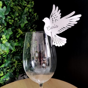 Puede incluir: Una tarjeta de lugar en forma de paloma blanca, con el nombre "Carla" en escritura cursiva, se posa sobre una copa de vino. La paloma tiene recortes detallados en las alas. Fondo negro con follaje verde a la izquierda.