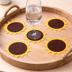May include: A wooden serving tray with rope handles holds a wine glass and five sunflower-shaped coasters. The coasters have a yellow petal design around a brown center. The tray and coasters are on a light-colored wooden surface.