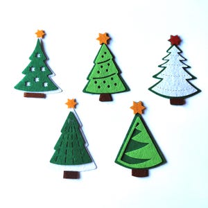 Puede incluir: Cinco adornos de árbol de Navidad de fieltro en varios diseños. Dos árboles verdes con detalles blancos, un árbol blanco con detalles verdes y dos árboles verdes con detalles de estrellas amarillas.