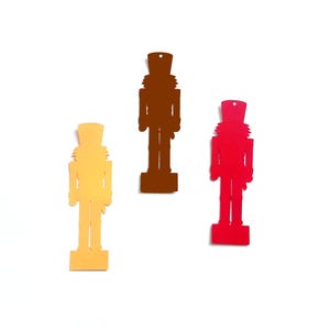 Op de afbeelding: Drie kleurrijke notenkraker ornamenten in goud, bruin en rood. Elk ornament is een uitgesneden silhouet van een traditionele notenkraker soldaat, met een gat aan de bovenkant om op te hangen. De ornamenten staan tegen een witte achtergrond.