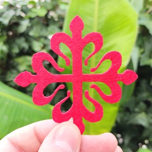 Puede incluir: Un recorte de fieltro rojo brillante con forma de cruz estilizada y cuatro brazos curvos simétricos. El diseño presenta puntas en los extremos superior, inferior, izquierdo y derecho, con intrincados recortes redondeados entre los brazos.