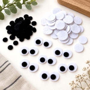 May include: A collection of craft supplies including a pile of small black felt circles, a pile of white felt circles, and several pairs of white felt circles with black centers designed to resemble eyes, arranged on a plain white surface.
