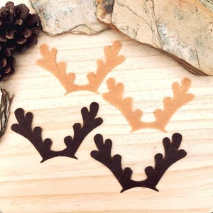 May include: Four felt reindeer antlers in shades of brown and tan are arranged on a wooden surface. Two are dark brown, and two are tan. A pine cone and stone fragments are in the background.