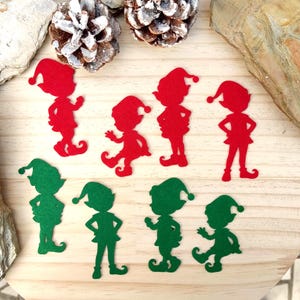 以下が含まれることがあります： さまざまなポーズの赤と緑のフェルトエルフの切り抜き。切り抜きは明るい木材の表面に配置され、背景には松ぼっくりと木片があります。エルフは尖った帽子と靴を履いています。
