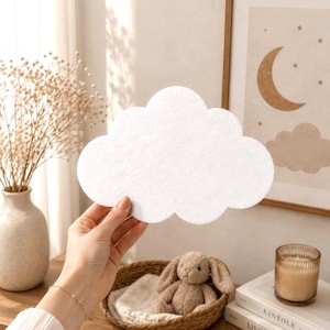 Puede incluir: Un recorte blanco con forma de nube hecho de un material similar al fieltro con una textura suave y mate. El objeto presenta una silueta clásica, redondeada y de múltiples lóbulos, que representa una forma de nube estilizada sin patrones.