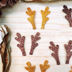 Op de afbeelding: Close-up van glitterende geweivormen in goud en bordeaux. De geweien zijn gerangschikt op een licht houten oppervlak, wat wijst op een feestelijk of vakantiethema. Geschikt voor knutselen of decoratie.