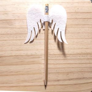May include: A wooden pencil adorned with a white felt angel wing design. The wings feature intricate feather detailing. The pencil is positioned centrally on a light brown wooden surface.