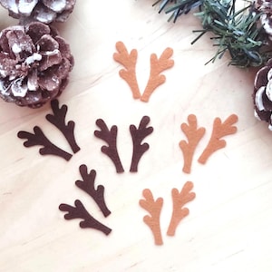 Puede incluir: Un conjunto de cuernos de reno de fieltro en varios tonos de marrón y beige, dispuestos sobre una superficie de madera clara. Los cuernos están en pares, con piñas y ramitas de hoja perenne en el fondo. Una imagen festiva y artesanal.