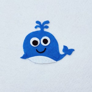 Pode incluir: Uma baleia azul de feltro com olhos brancos e barriga branca. A baleia tem uma boca sorridente.